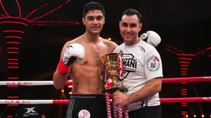 A boxer in gloves and a coach smiling together with a trophy in the boxing ring.