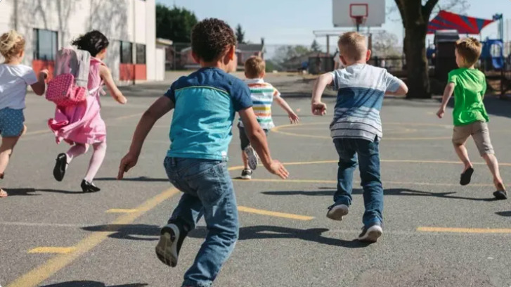 A group of children running on a playground, with a basketball hoop and colorful equipment in the background.