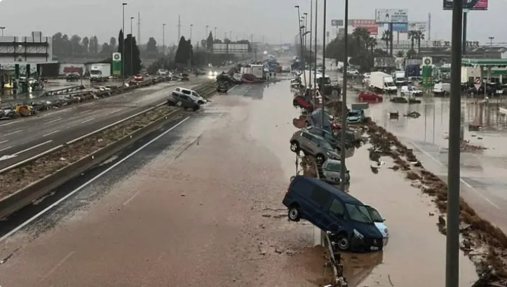 A flooded highway with abandoned cars, including one tilted off the road, and trucks in the distance.