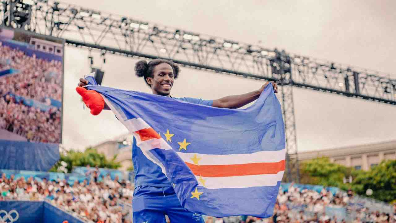 An Olympian smiles while holding the Cape Verde flag, with a crowd and a large screen in the background.