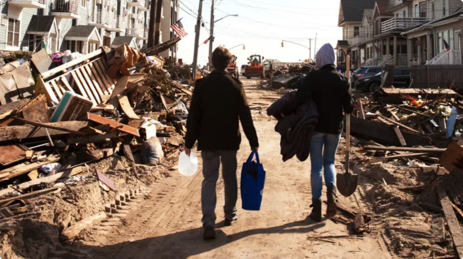 Two people walking through a disaster-stricken street with debris, carrying supplies and walking past damaged homes.