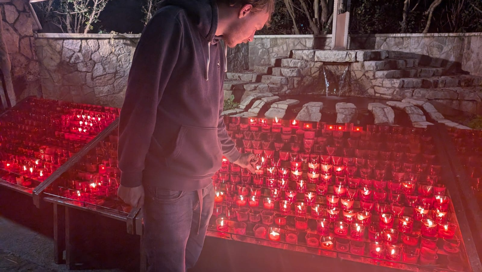 A person in a hoodie lights candles in front of a large arrangement of red candles at night.