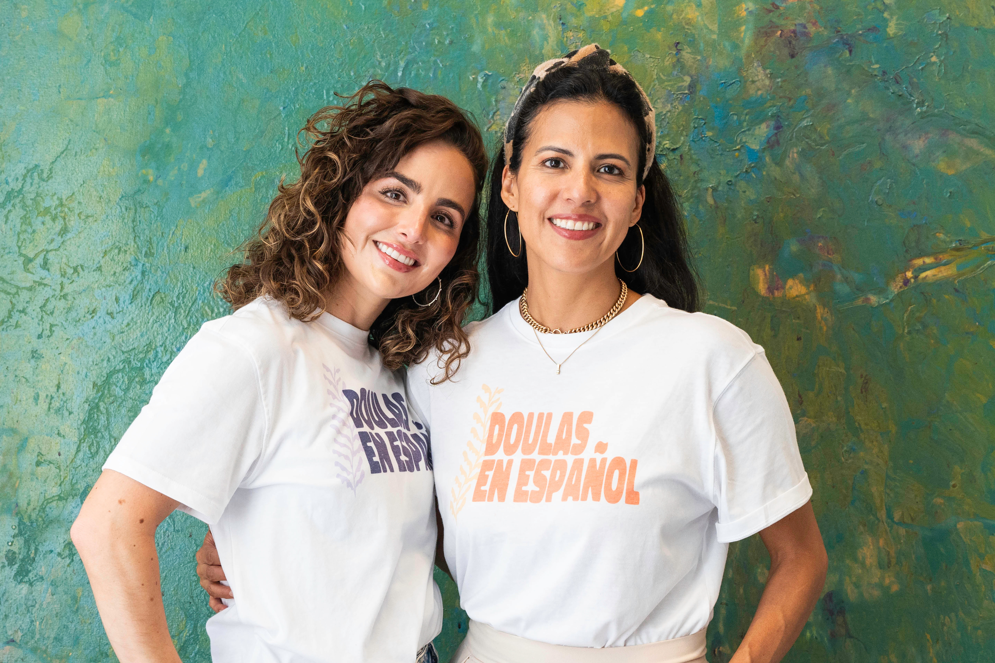 Two women standing together, smiling and wearing 'Doulas en Español' t-shirts in front of a colorful background.