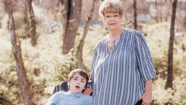 A woman standing beside a child in a wheelchair, both smiling outdoors in a sunny setting with trees in the background.