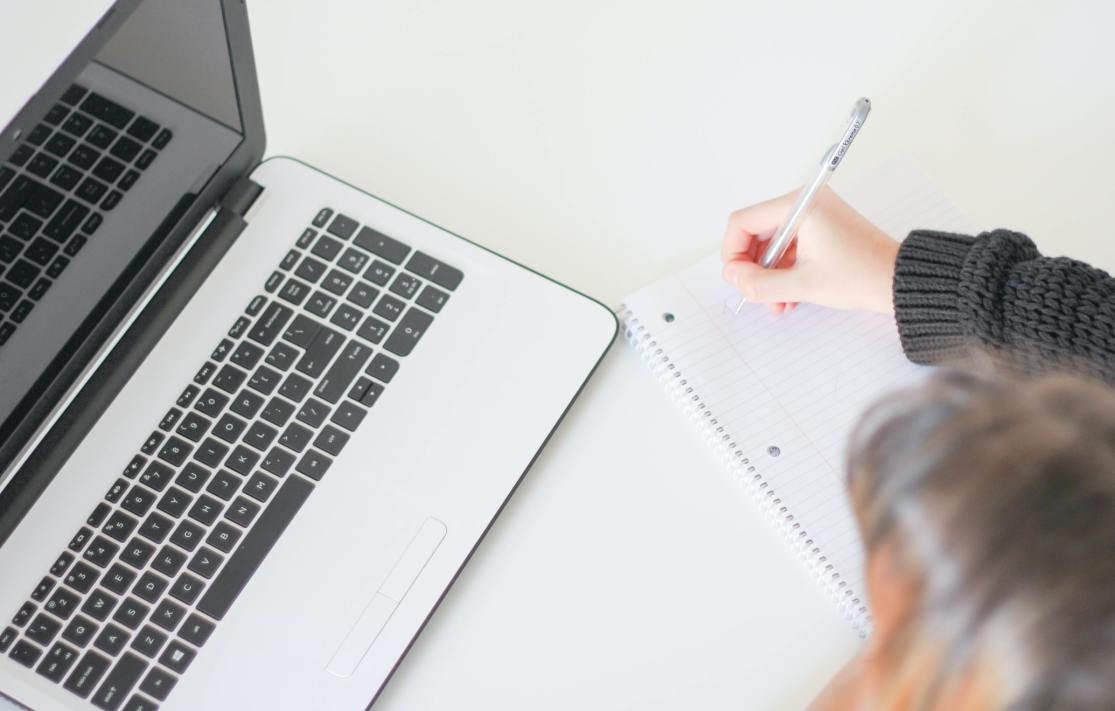 A person writing in a notebook next to a laptop